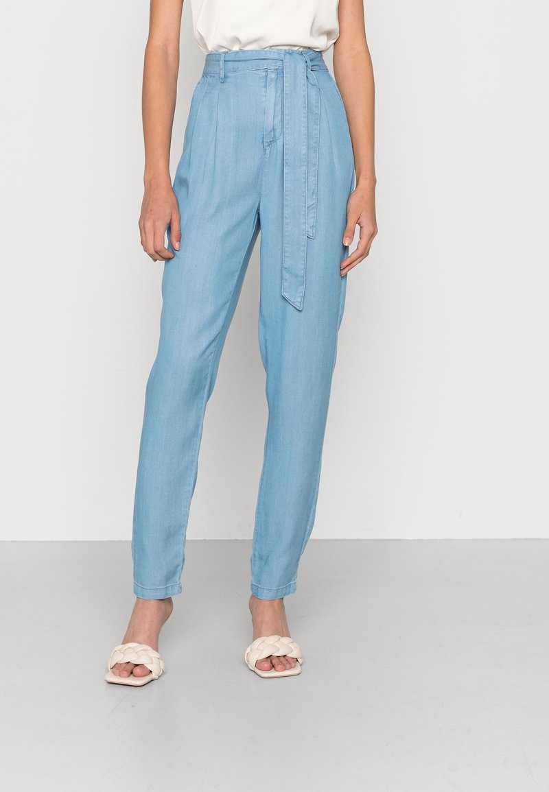 Femme portant un pantalon taille haute bleu clair à taille fuselée avec une ceinture assortie en tissu, un haut blanc sans manches et des sandales blanches tressées à bout ouvert.