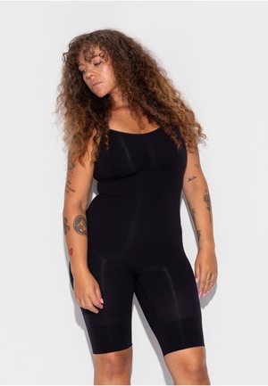 Frau mit lockigem Haar, die einen enganliegenden schwarzen ärmellosen Body trägt, steht vor einem einfarbigen weißen Hintergrund mit Tätowierungen an beiden Armen.