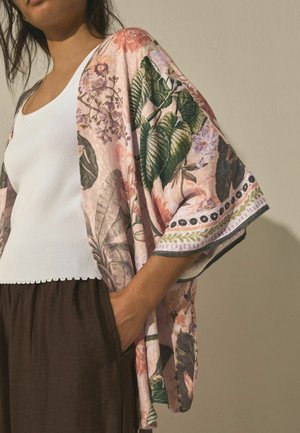 Woman wearing a white ribbed tank top, brown pants, and a loose pink kimono with large green and purple floral prints.