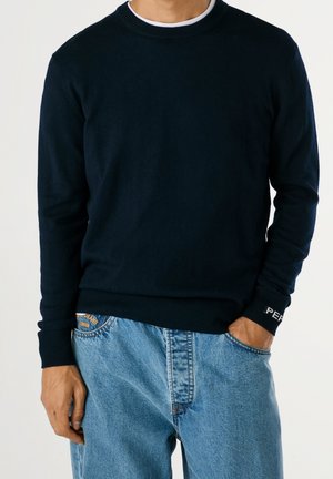 Mann trägt dunkelblauen Pullover über weißem Hemd und lockere hellblaue Jeans, eine Hand in der Tasche, vor einfarbigem Hintergrund.