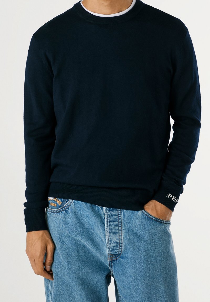 Homme portant un pull bleu marine foncé par-dessus une chemise blanche et un jean bleu clair ample, une main dans la poche, sur fond uni.