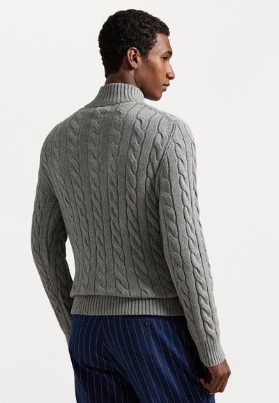 Suéter cinza em tricot com gola e bainha caneladas, apresentando padrões elevados e um acabamento texturizado. Usado com calças azul-marinho com riscas.