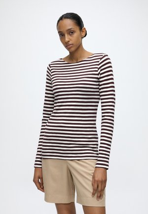 Femme aux cheveux foncés attachés en chignon, portant un haut à manches longues rayé bordeaux et blanc et un short beige longueur genou, debout devant un fond uni.