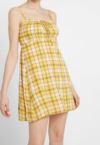 Keltainen ruudullinen mekko ohuiden hihnojen kanssa, kerätty yläosa ja kiristysnyöri. Sisältää vuorotellen valkoisia ja mustia ruutukuviota.