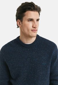 Giovane uomo con capelli scuri che indossa un maglione a maglia blu navy con granuli, che guarda di lato contro uno sfondo chiaro e semplice.