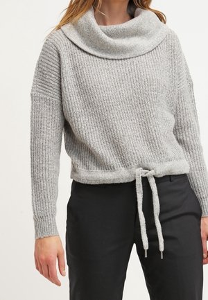 Femme portant un pull en maille gris avec un col bénitier et une taille à cordon, associé à un pantalon noir sur un fond uni.