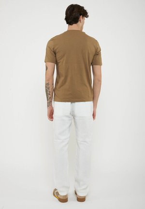 Man met een bloementatoeage, die een bruin shirt met korte mouwen, witte broek en bruine schoenen draagt, staat met zijn rug naar de camera tegen een effen achtergrond.