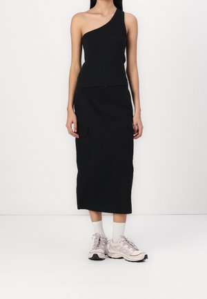 Haut noir asymétrique à une épaule associé à une longue jupe noire avec poches cargo, porté avec des baskets rose clair et des chaussettes blanches.