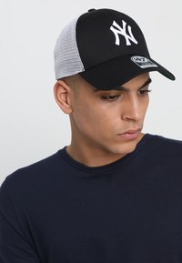 Căciulă de baseball neagră cu logo alb brodat și spate din plasă, având o parte inferioară verde a vizorului și sistem de prindere tip snapback.