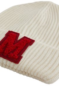 Kremas spalvos megzta kepurė su ribota tekstūra; priekyje didelis raudonas medvilnės pleistras su „M“ raide.