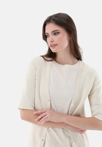 Strickcardigan in hellem Beige mit kurzen Ärmeln, mit einem lockeren Schnitt und Knopfverschluss. Über einem passenden T-Shirt getragen.