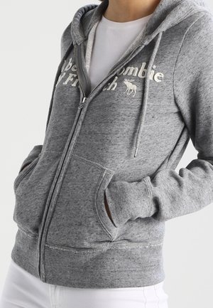 Person iført en grå hættetrøje med lynlås, forlommer og hvid tekstlogo, over en hvid T-shirt og hvide bukser.