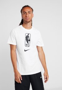 Camiseta blanca de Nike con un gráfico del logo de la NBA en negro centrado en el pecho, con mangas cortas y un escote redondo.