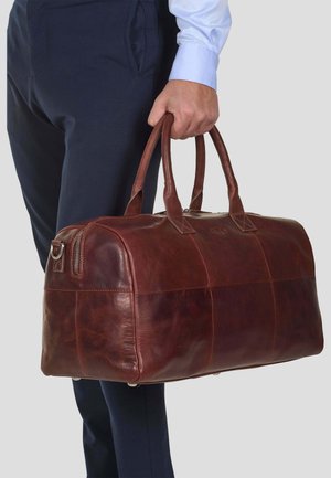 Brown leather duffel bag with dual handles, rounded shape, distinct seams, and silver hardware accents. Bag is being held by a person.
