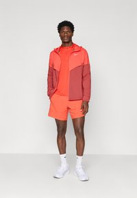 Nike αθλητική ενδυμασία που περιλαμβάνει ένα κόκκινο μπουφάν με κουκούλα και ταιριαστές κόκκινες βερμούδες. Το ύφασμα φαίνεται ελαφρύ με λεία υφή.