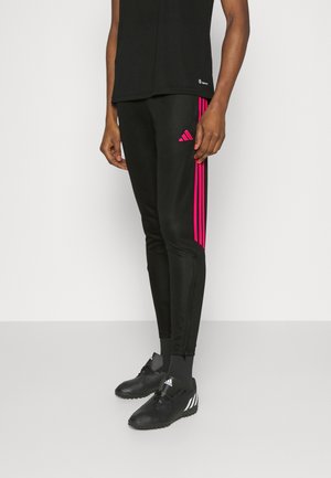 Osoba ubrana w czarne sportowe spodnie z różowymi paskami po bokach i czarne buty sportowe stoi na białym tle.