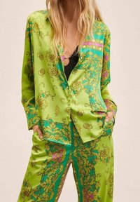Vrouw in een groen satijnen pak met sierlijke gouden en turquoise bloempatronen, handen in de zakken, zwarte top zichtbaar onder het pak.