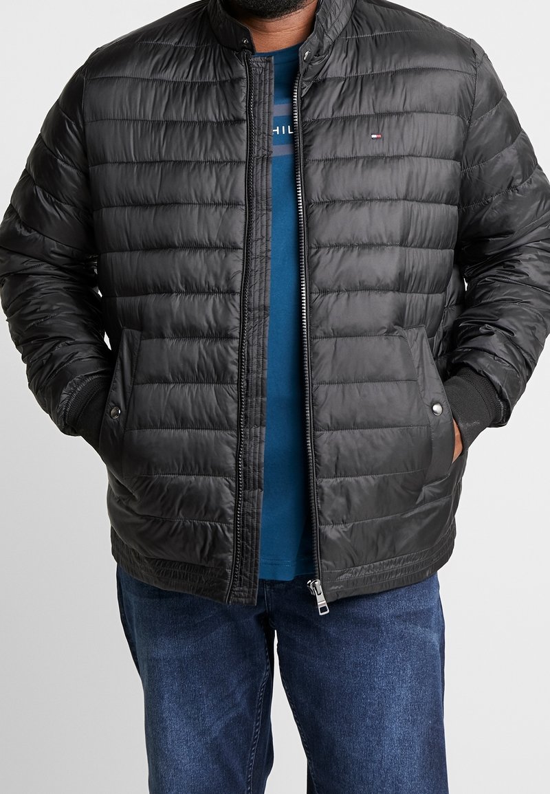 Veste rembourrée noire matelassée avec un col montant, fermeture éclair, deux poches avant et des poignets élastiques. Le matériau semble lisse et léger.