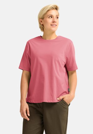 Vrouw met kort blond haar draagt een lichtroze shirt met korte mouwen en donkerolijfgroene broek, staat met één hand in de zak en kijkt naar rechts.