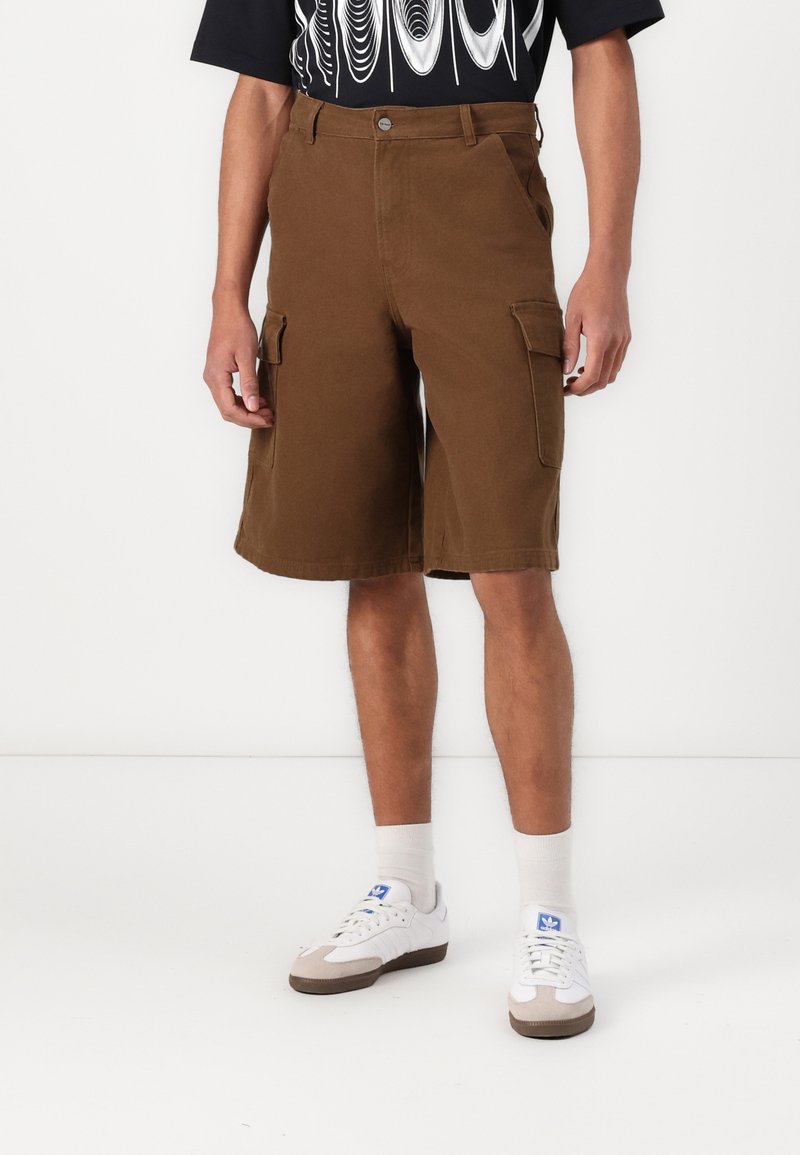 Pantalones cortos de carga marrones con dos bolsillos laterales, hechos de una tela duradera, con un diseño de corte relajado y pierna recta. Combinados con zapatillas blancas.