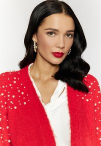 Cardigan rosso con decorazioni di perle, maglia a texture, indossato sopra una camicia bianca. La modella ha i capelli lunghi e scuri e indossa orecchini a cerchio.