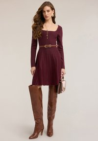 Bordeauxrotes, geripptes Strickkleid mit langen Ärmeln, tailliertem Schnitt und ausgestelltem Rock. Brauner Ledergürtel und kniehohe braune Stiefel. Rosa Handtasche.