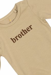 Beige heldragt med lange ærmer lavet af blødt stof med brunt printet tekst "brother". Inkluderer trykknapper ved skulderen for nem påklædning.