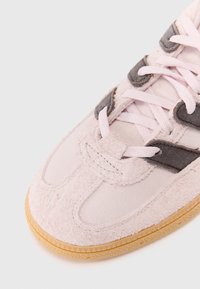 Κοντινό πλάνο της μύτης ενός ελαφριά ροζ σουέτ αθλητικού παπουτσιού με υφή σόλας και ανοιχτό ροζ κορδόνια που περνούν μέσα από σκούρους καφέ θηλιές.