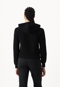 Schwarze Strickjacke mit gerippter Textur, ausgestattet mit einer Kapuze und langen Ärmeln. Getragen mit dunklen Jeans, von hinten betrachtet.