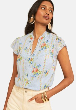 Kvinde iført lyseblå blomsterbluse med blondekant, cremefarvede bukser, store runde øreringe og to gyldne armbånd på venstre håndled.