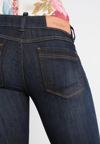 Dark blue denim jeans feature a mid-rise waist, three pockets, and a tan leather tag with "Marc O'Polo" stitched on the back.