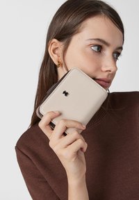 Beige textured wallet with dark brown accents, featuring a small black logo. The wallet is held in a hand against a neutral background.