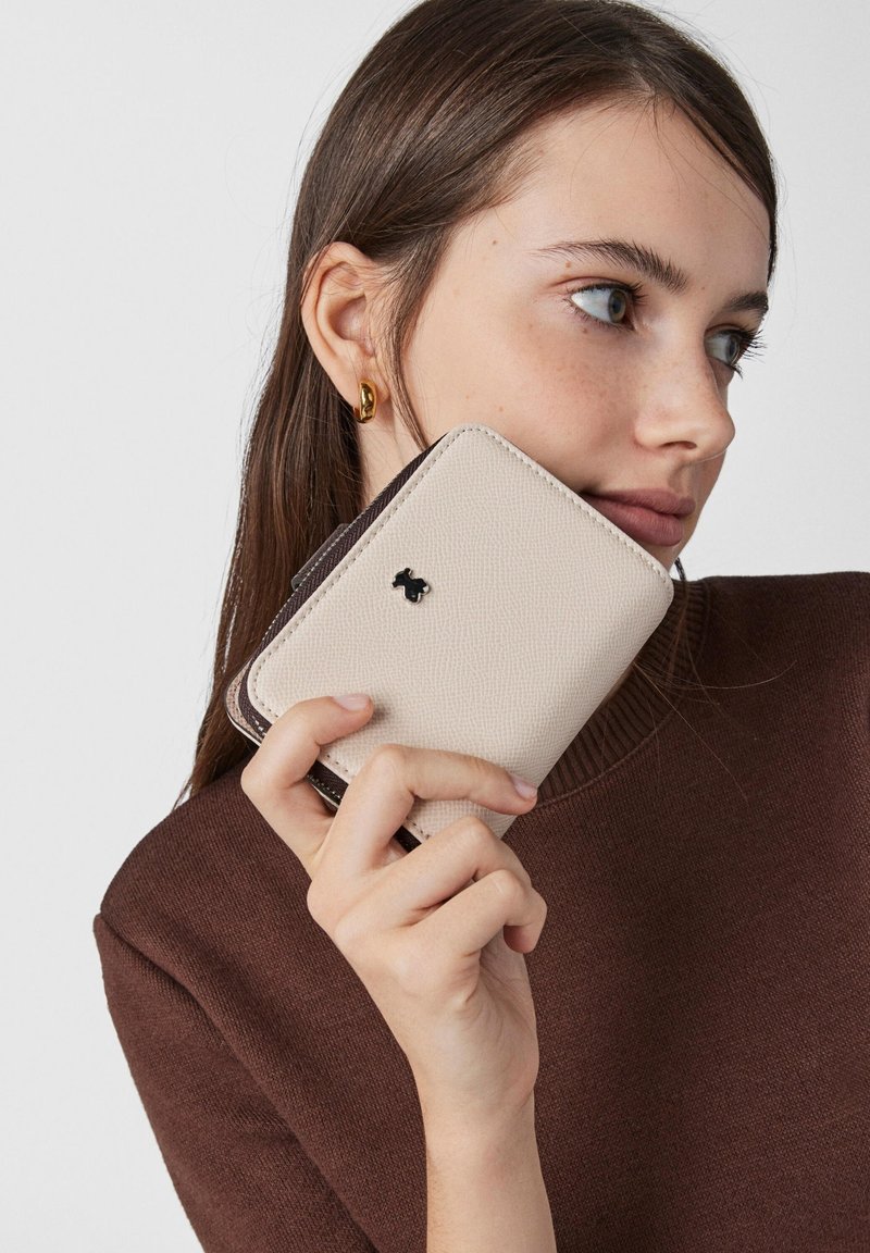 Beige textured wallet with dark brown accents, featuring a small black logo. The wallet is held in a hand against a neutral background.