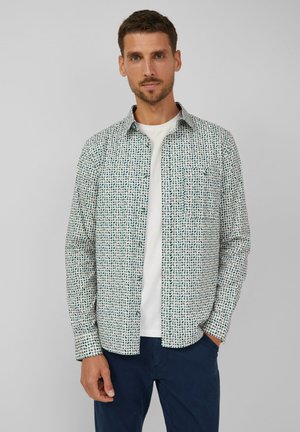 Man in een wit met patroon bedrukte overhemd over een wit T-shirt met een marineblauwe broek, staand tegen een effen lichtgrijze achtergrond.