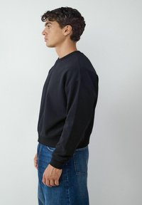 Czarna bluza z okrągłym dekoltem, rękawami raglanowymi i ściągaczami; połączona z niebieskimi dżinsami, przedstawiona z boku.