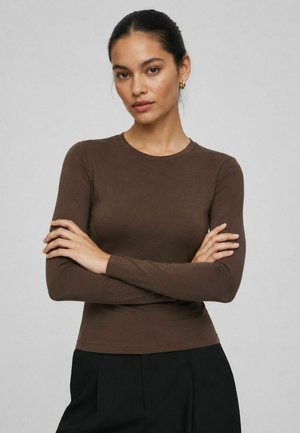 Femme aux cheveux foncés en chignon portant des boucles d'oreilles créoles dorées, un haut marron moulant à manches longues et un pantalon noir, les bras croisés.