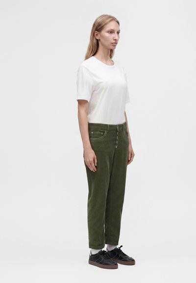 Femme debout portant un t-shirt blanc uni à manches courtes, un pantalon taille haute vert olive avec des boutons, des chaussettes blanches et des baskets noires.