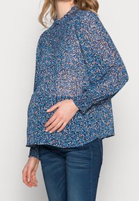Gravid kvinna som bär en blå blommig blus med långa ärmar och mörka jeans, som försiktigt håller om sin mage med ena handen.