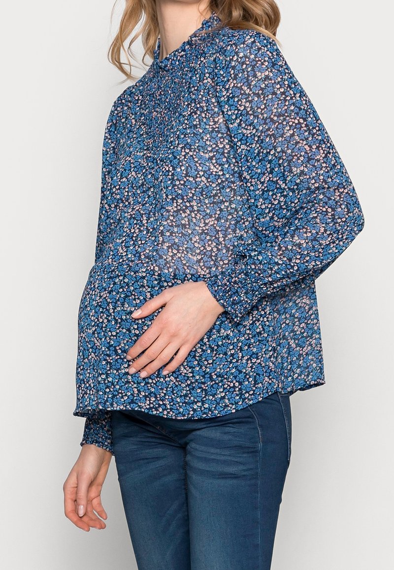 Gravid kvinna som bär en blå blommig blus med långa ärmar och mörka jeans, som försiktigt håller om sin mage med ena handen.