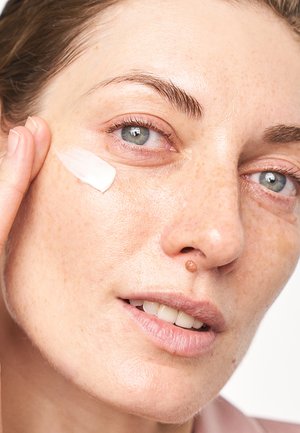 Nahaufnahme des Gesichts einer Person, die eine Creme mit glatter Textur auf ihre Wange aufträgt. Helle Hautfarbe mit Sommersprossen und sichtbaren Poren, blaue Augen.