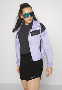 Casaco curto lilás com detalhes em preto, combinado com uma t-shirt preta e calções pretos. Os acessórios incluem óculos de sol grandes e refletivos.