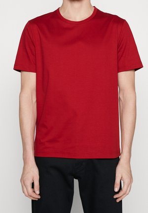 Mężczyzna ubrany w prostą, czerwoną koszulkę z krótkim rękawem i dekoltem w łódkę oraz czarne spodnie, stojący na jednolitym jasnym tle.