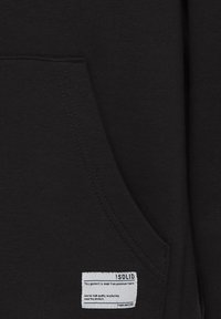 Sudadera negra con bolsillo tipo canguro, fabricada con un tejido de algodón texturizado y con una etiqueta de cuidado blanca cosida en la parte frontal.