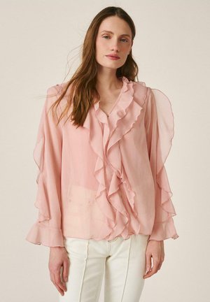 Mujer con cabello largo y castaño, vestida con una blusa rosa pálido translúcida con volantes en la parte delantera y las mangas, combinada con pantalones color blanco roto, de pie sobre un fondo neutro.