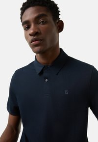 Granatowa koszulka polo wykonana z fakturowanej tkaniny. Posiada klasyczny kołnierzyk, rozpinany na trzy guziki oraz subtelne logo na piersi.