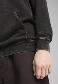 Felpa nera con una texture morbida, dotata di una tasca frontale e cuciture a contrasto. Indossata con pantaloni marrone scuro, mano appoggiata in modo casuale.