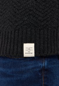 Schwarzer gestrickter Pullover mit einem strukturierten Fischgrätenmuster, der einen gerippten Saum und ein Stoffetikett mit der Aufschrift "True Denim for True People" aufweist.