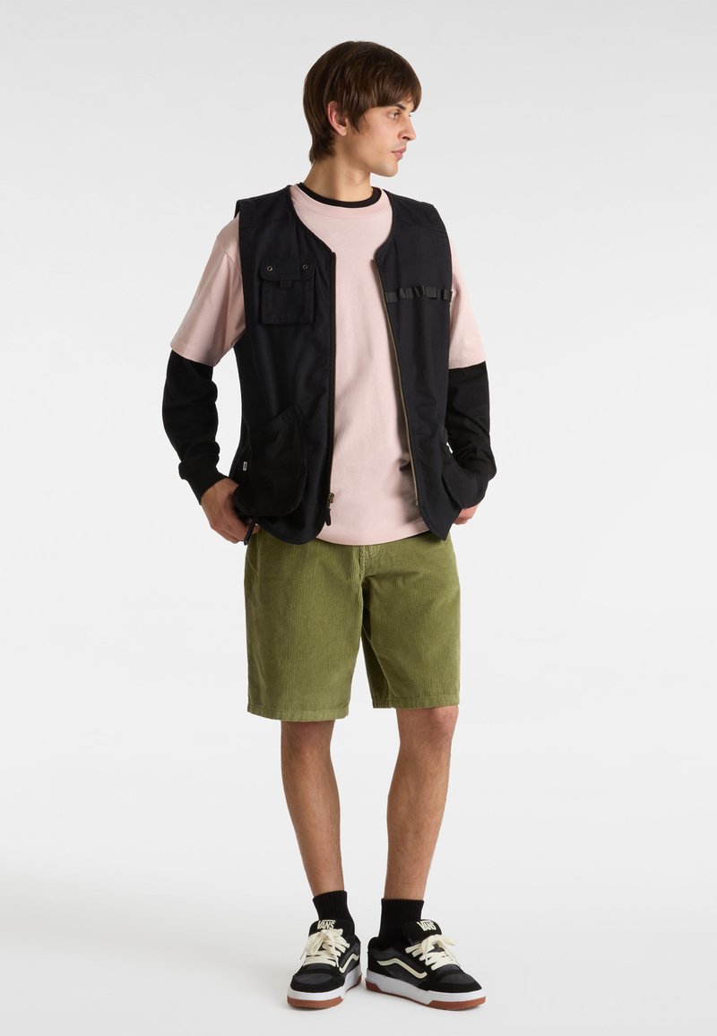 Svart ärmlös väst med framfickor över en rosa skjorta, klädd med gröna kordbyxor och svarta sneakers med vita snören.