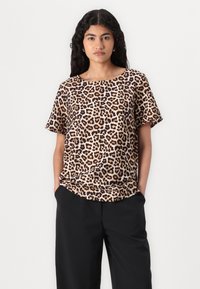 Korte erm blouse med leopardmønster i tan og brun, med bred halsudskæring og afslappet pasform, parret med sorte bukser. Glat stoftekstur.