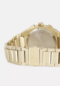 Kuldse tooniga käekell metalliga. Omab tekstureeritud pinda, ristkülikukujulisi linke ja ümmargust korpust, millel on nähtav kroon ja logo kinnitusel.